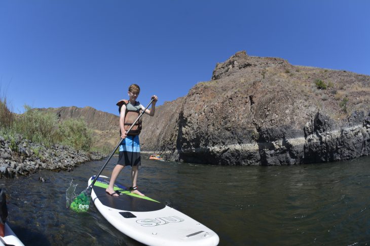 Kid in life vest standing on a paddle board on the John Day River with mountains behind him
