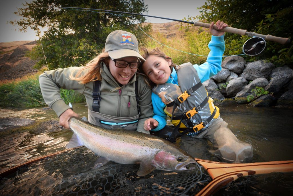 Guide, Mia, and little girl with fishing rod sitting in river pulling a steelhead out of the water