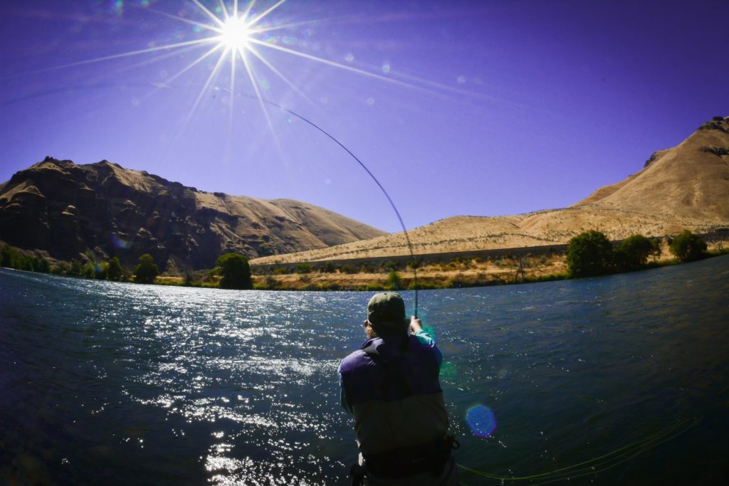 Person casting fishing line with river, mountains, and a sunny sky in front of them