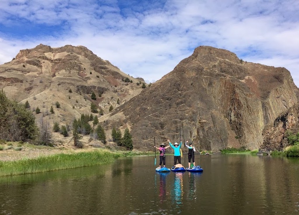 Three people in a row standing on paddle boards on the John Day River with mountains in the background