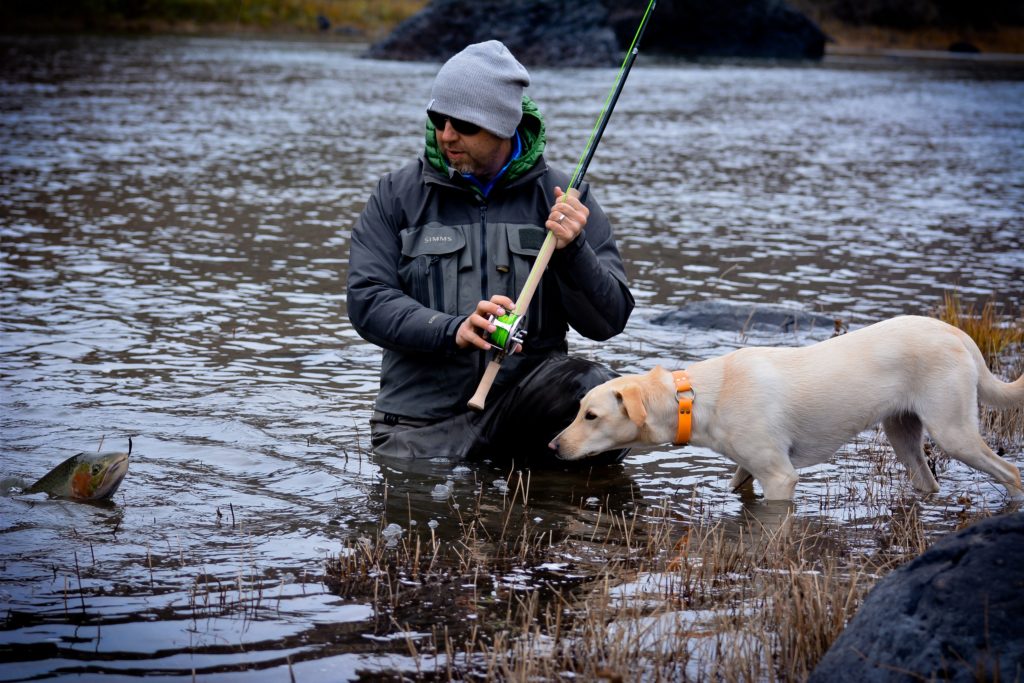 Man in fishing gear and beanie standing in the river holding a fishing rod with a dog beside him staring at the fish