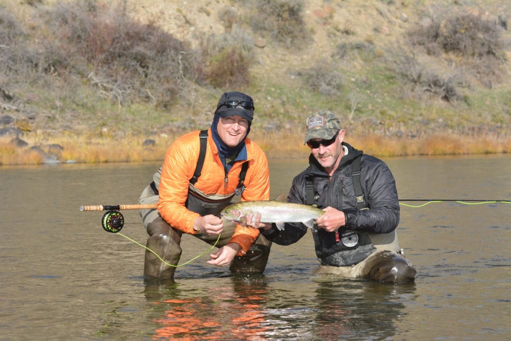 Two men in fishing gear crouching in the water holding a fishing rod and a steelhead fish