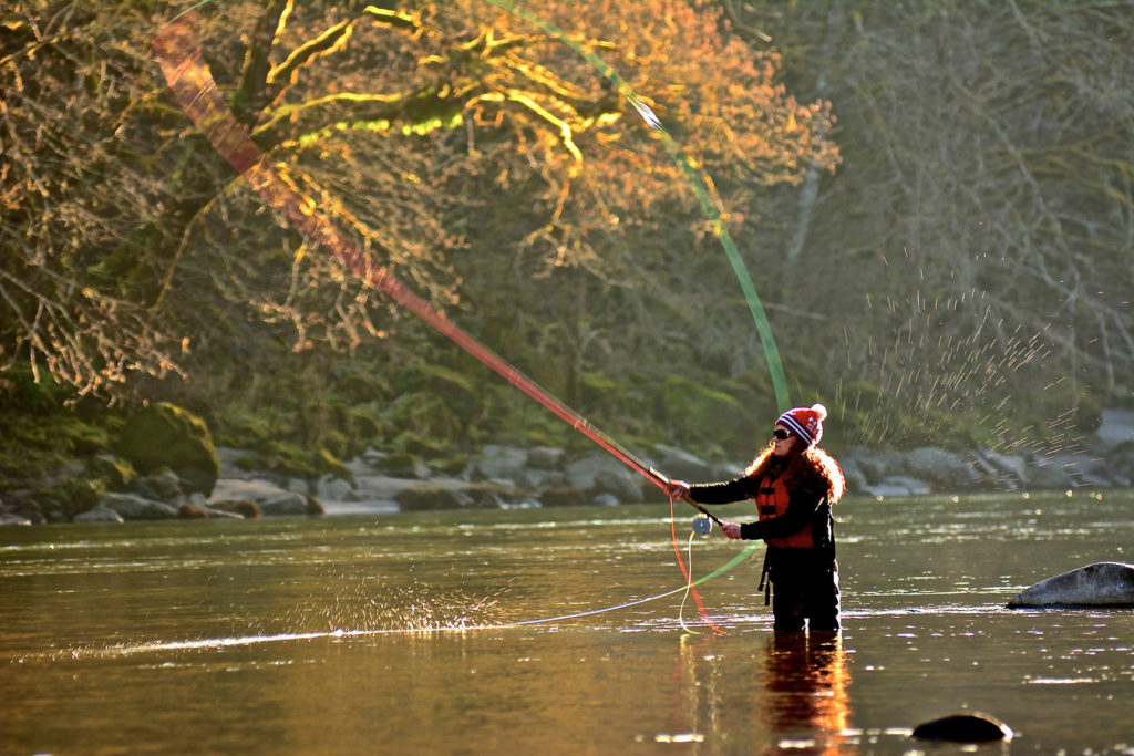 Woman in fishing gear and life vest casting a fly fishing line into the Sandy River on a sunny day