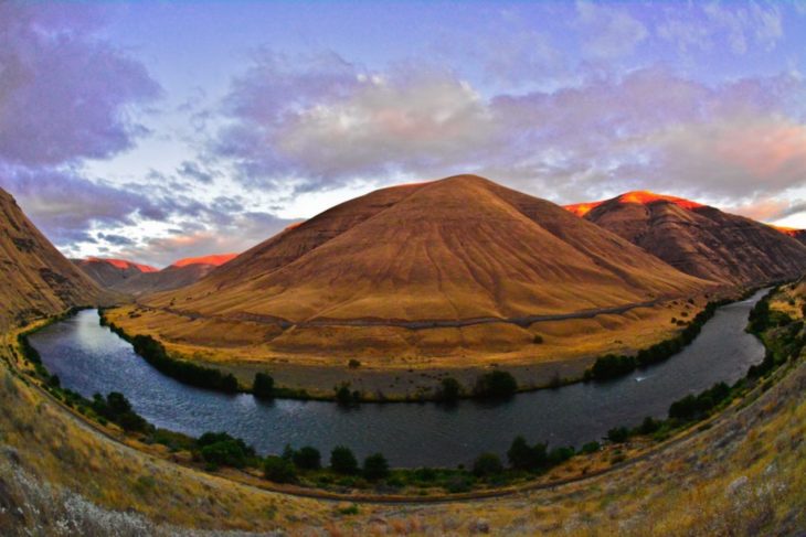Large mountain with Deschutes river wrapping around it and an orange and purple sunset in the sky